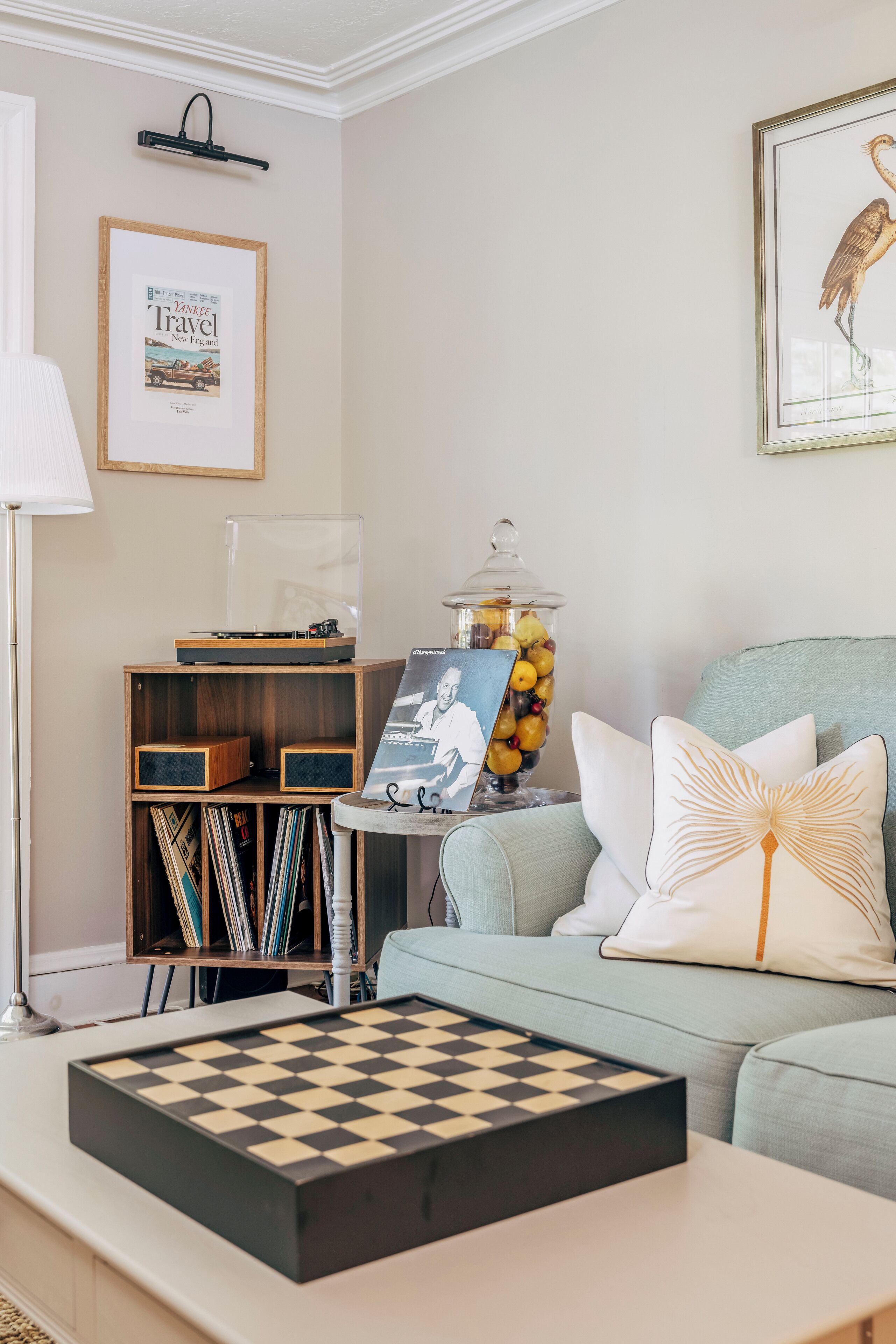 A living room with a light blue sofa, a coffee table with a chessboard, a bookshelf with vinyl records, a framed travel poster, and a fruit jar.
