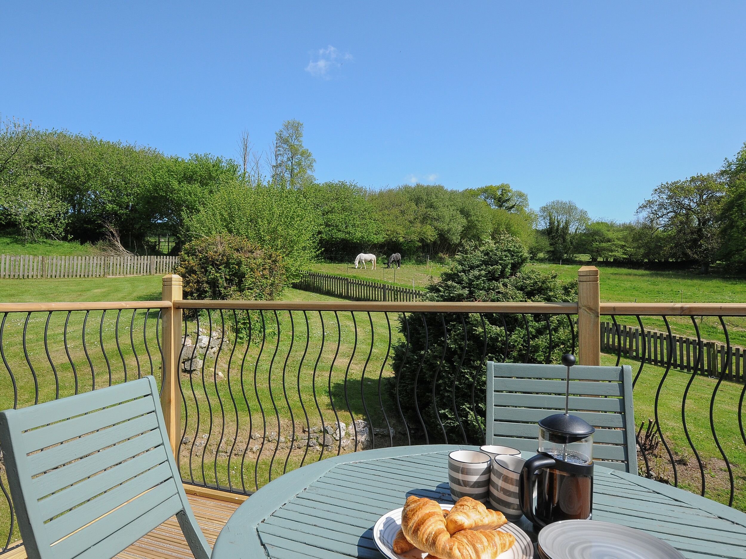 Una mesa con croissants y una cafetera, con vistas a un jardín cercado con caballos.
