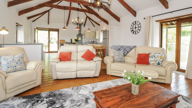 A spacious living room with high ceilings and exposed wooden beams, featuring two cream-colored sofas with patterned cushions, a wooden coffee table, and a chandelier. - Mill House (Lostwithiel)