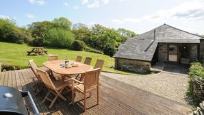 A wooden deck with a round table and chairs, a barbecue grill, and a stone building with a sloped roof. - Mill House (Lostwithiel)