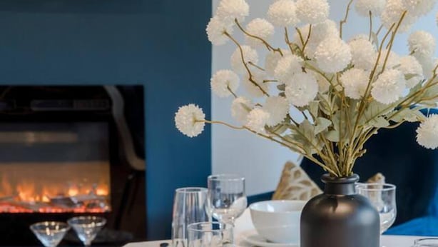 Mesa de comedor dispuesta con platos blancos, tazones y vasos transparentes, un florero con flores blancas y una chimenea al fondo.