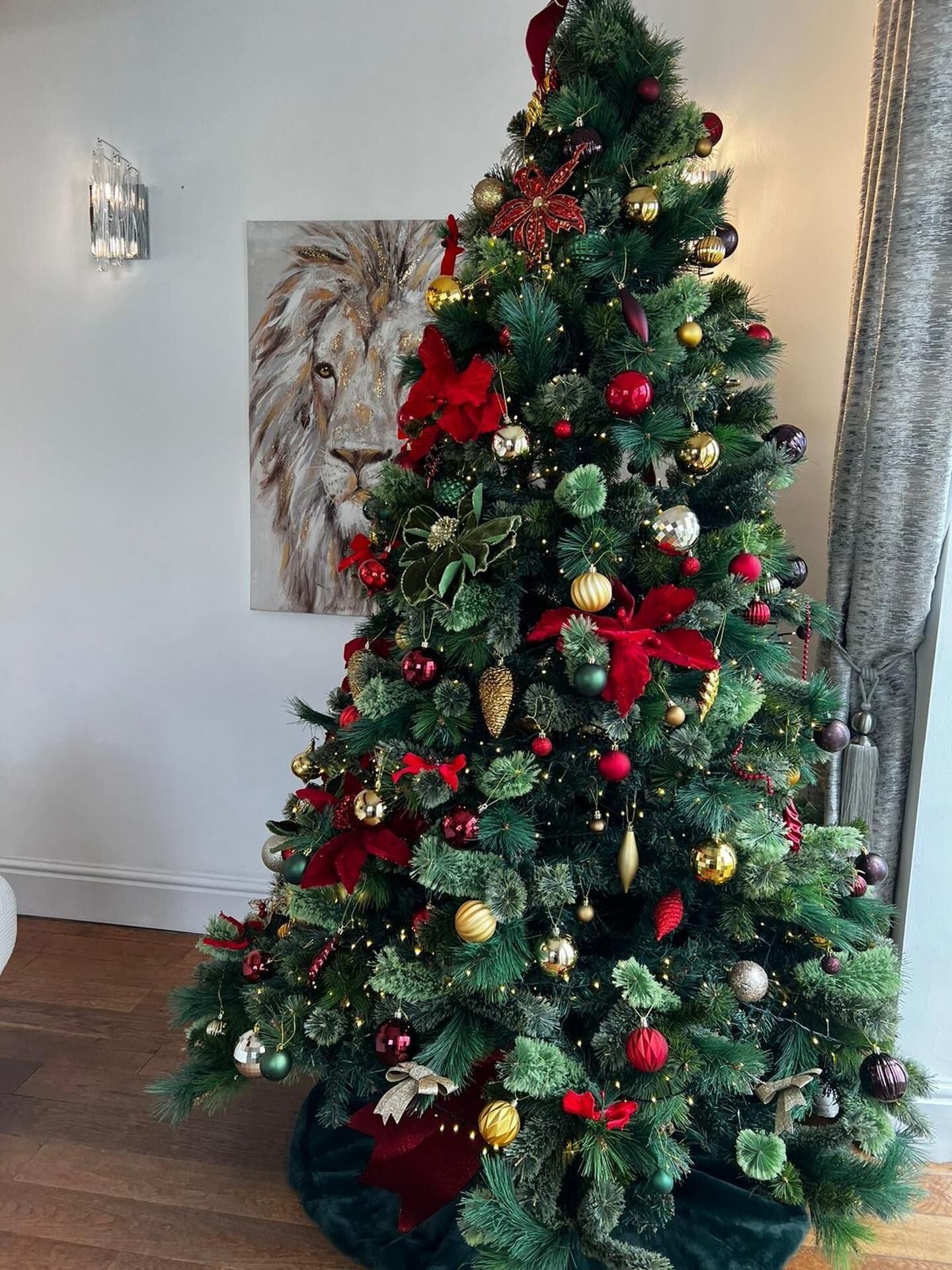 Ein Weihnachtsbaum mit verschiedenen Dekorationen und Lichterketten, platziert in einem Raum mit einem gerahmten Bild an der Wand.