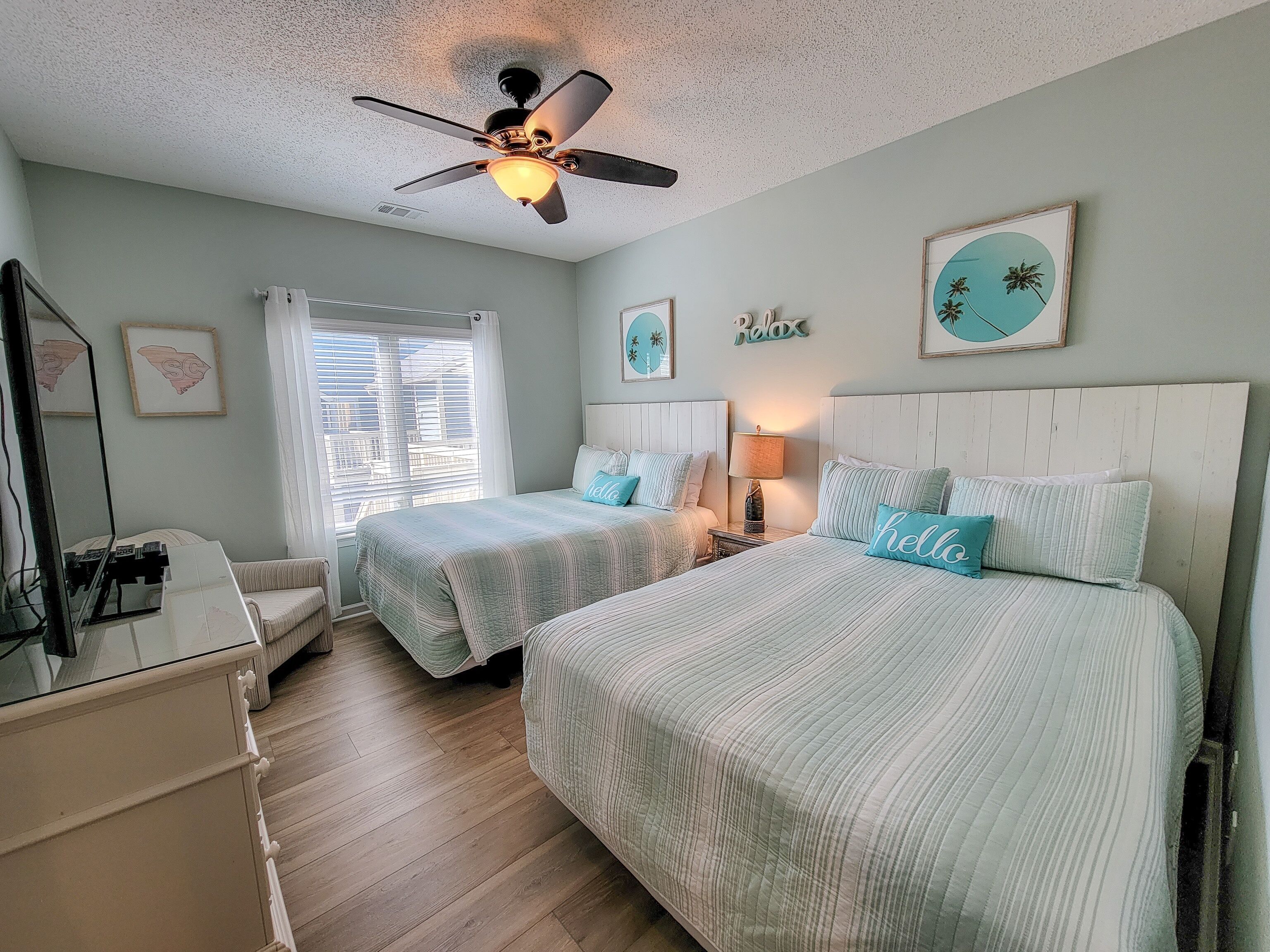 A bedroom with two beds, a ceiling fan, a window with blinds, and a desk with a lamp.