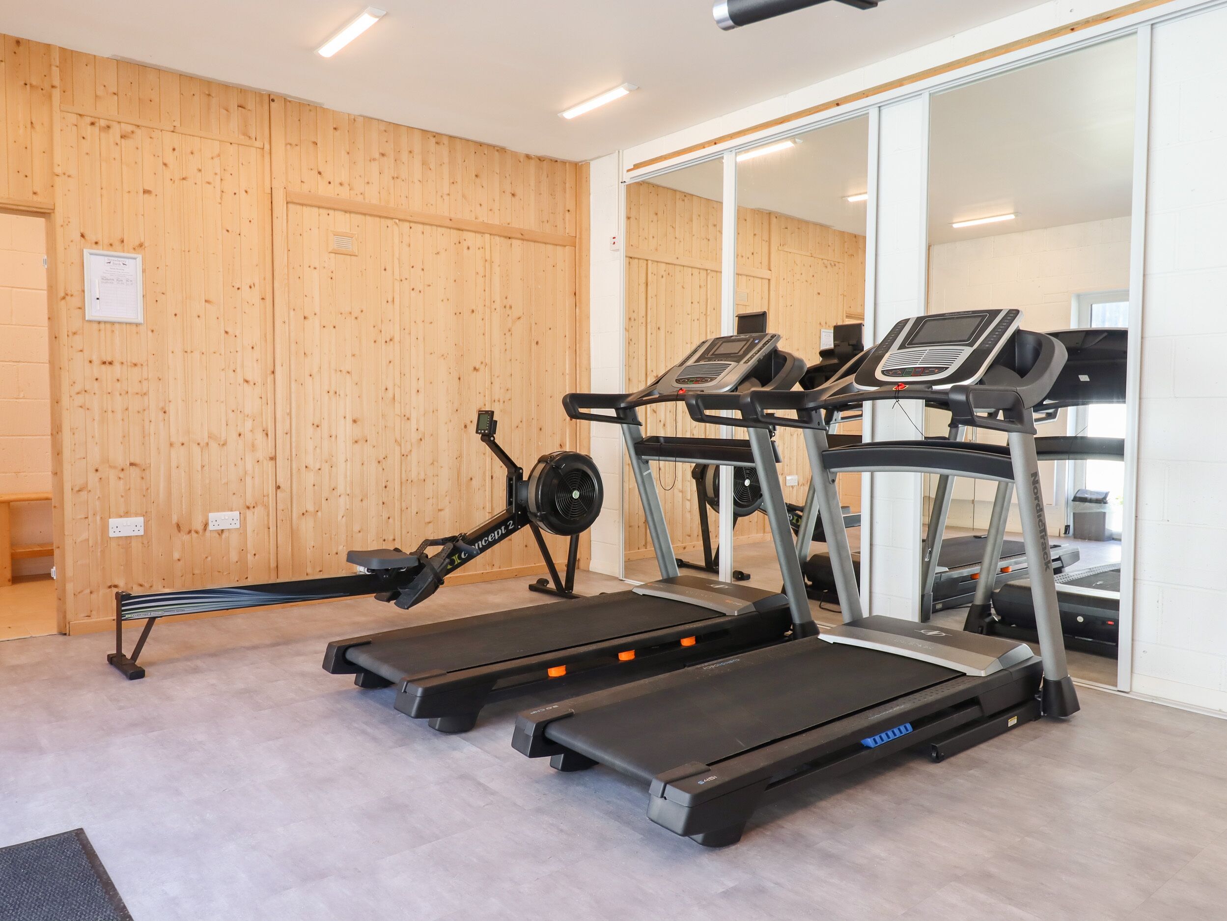 Un gimnasio con dos cintas de correr, una máquina de remo y una pared de espejos.
