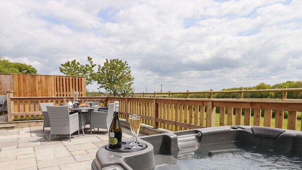 A hot tub with a bottle of wine and glasses on a table, surrounded by wicker chairs and a wooden deck. - Strawberry Bank - Kingfisher (Holsworthy)