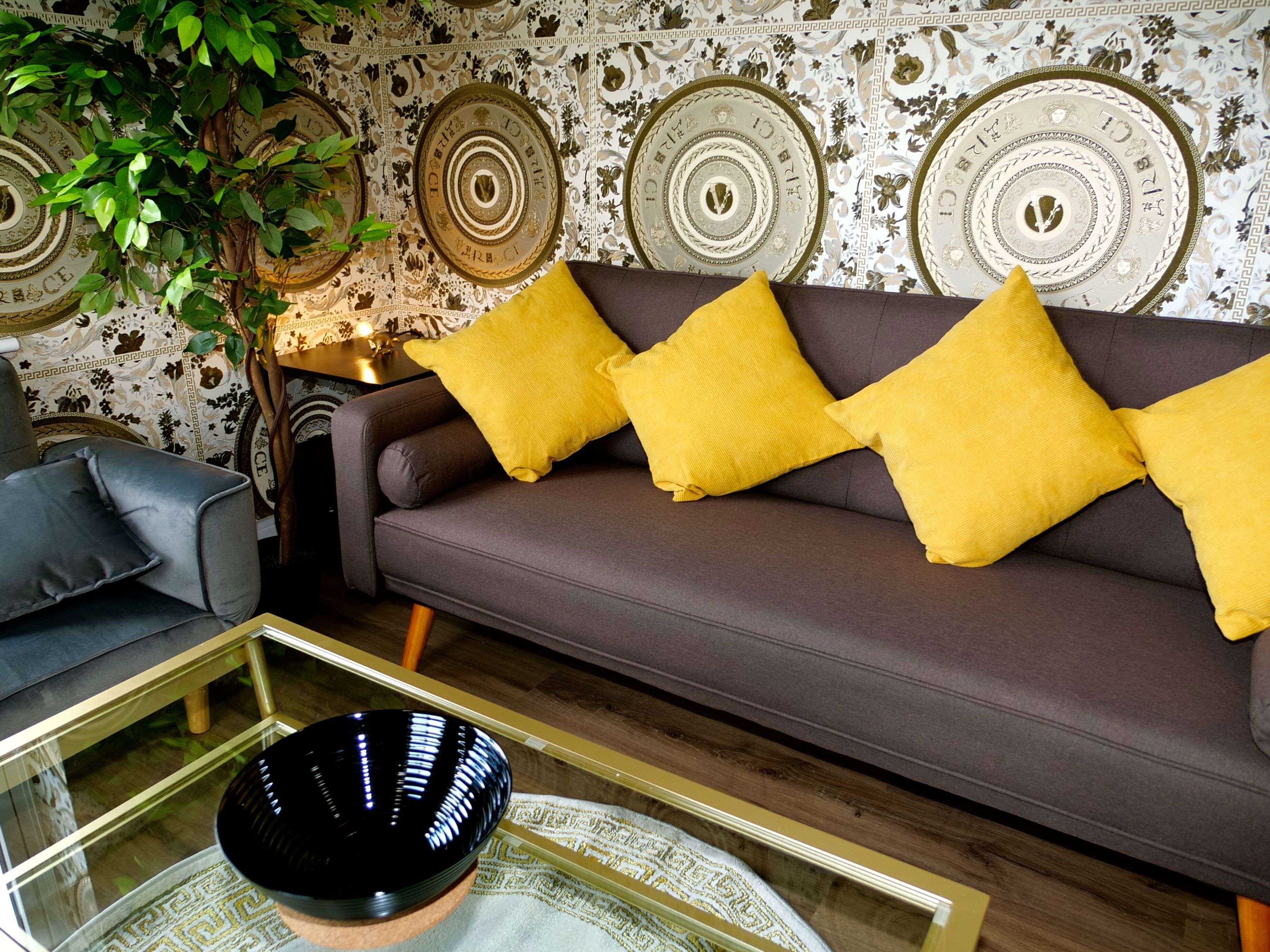 Una sala de estar con un sofá marrón, cojines amarillos, una mesa de centro de vidrio y una pared decorativa con patrones circulares.
