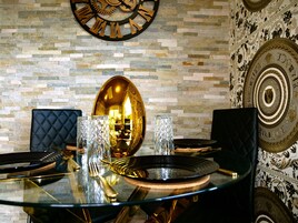 A dining table with a glass top, surrounded by black upholstered chairs, set with gold cutlery and crystal glasses, against a stone wall with a large clock and decorative plates.