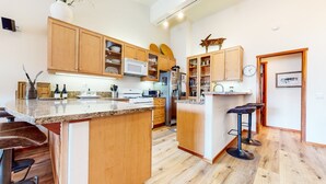 A modern kitchen with wooden cabinets, granite countertops, a built-in microwave, a stainless steel refrigerator, and a clock on the wall.