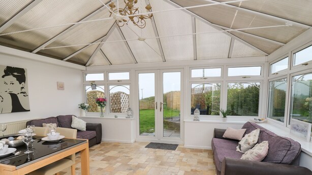 A conservatory with a tiled floor, two sofas, a dining table, and large windows offering a view of the garden. - Ty Cariad (Carmarthen)