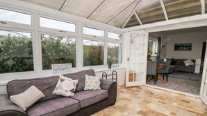 A conservatory with a grey sofa, a wooden chest, and a dining area with a grey sofa and a chair. - Ty Cariad (Carmarthen)