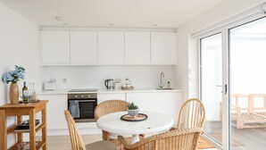 Ett modernt kök med vita skåp, ett matbord med flätade stolar och en skjutdörr i glas som leder ut till ett utomhusområde.
