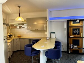 A compact kitchen with a central island, a microwave, a stove, and a sink. There are blue chairs around the island and a wooden bookshelf with books and decorative items. - Stunning View Seaside 1-bed Apartment in Saronida (Saronikos)