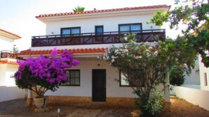 A two-story building with a balcony, a door, and a small garden with purple flowers.