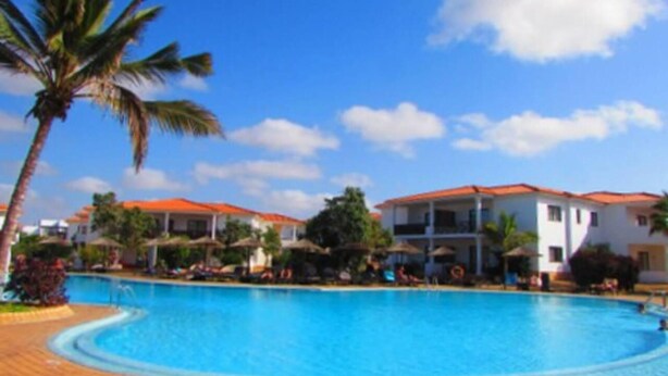 A swimming pool with a curved edge, surrounded by palm trees and buildings with red roofs.