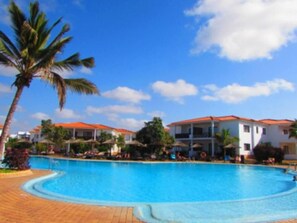 A swimming pool with a curved edge, surrounded by palm trees and buildings with red roofs. - Gorgeous 3-bed Villa Tortuga Beach Resort (Sal)