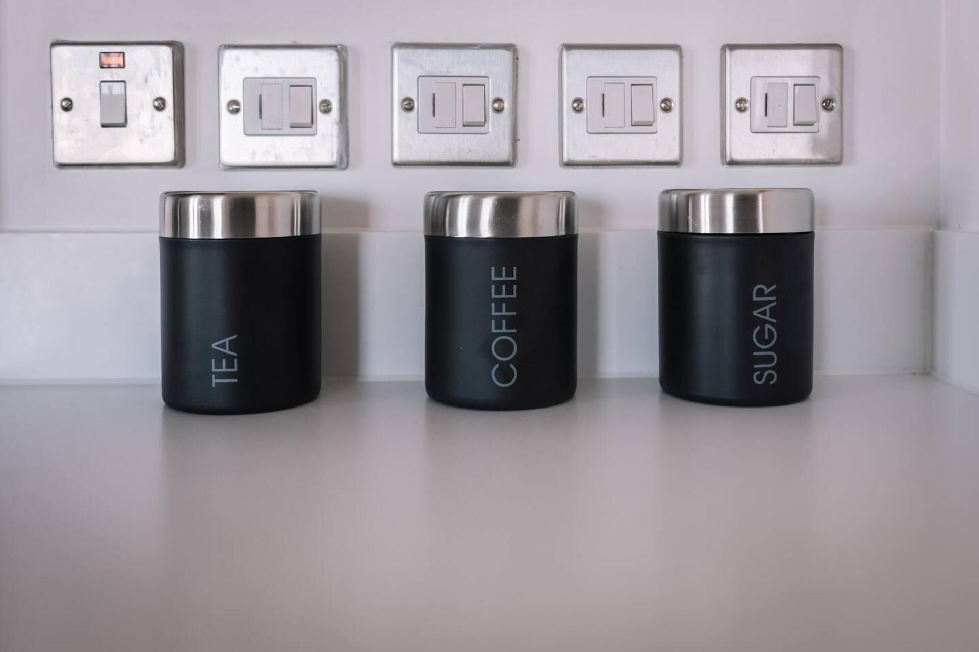 Three black containers labeled "TEA," "COFFEE," and "SUGAR" on a white shelf against a wall with five light switches above.
