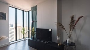 A modern living room with a large TV on a black entertainment unit, a small side table with a lamp, and a view of the city through floor-to-ceiling windows. - Birmingham Luxury Apartment - Free Wifi (Birmingham)