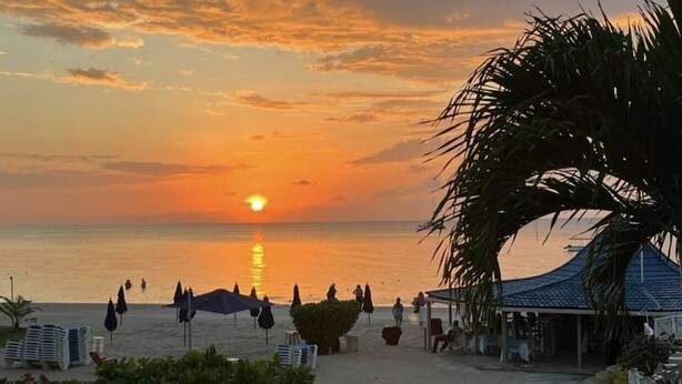 A beach scene with a sunset, palm trees, and beach umbrellas. - Seacret Studios - Cozy Jamaican Retreat Apts by RedAwning (Negril)