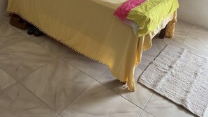 A bed with a yellow bedspread, a wooden chair, a window with curtains, and a wall-mounted shelf with decorative items. - Seacret Studios - Cozy Jamaican Retreat Apts by RedAwning (Negril)