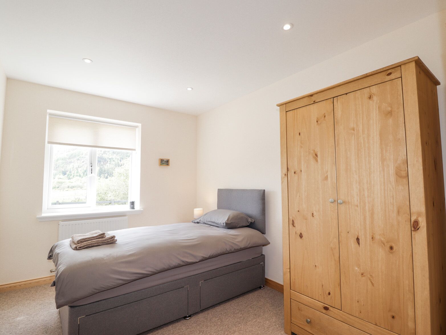 A bedroom with a bed, a wooden wardrobe, a radiator, and a window with a view of greenery.