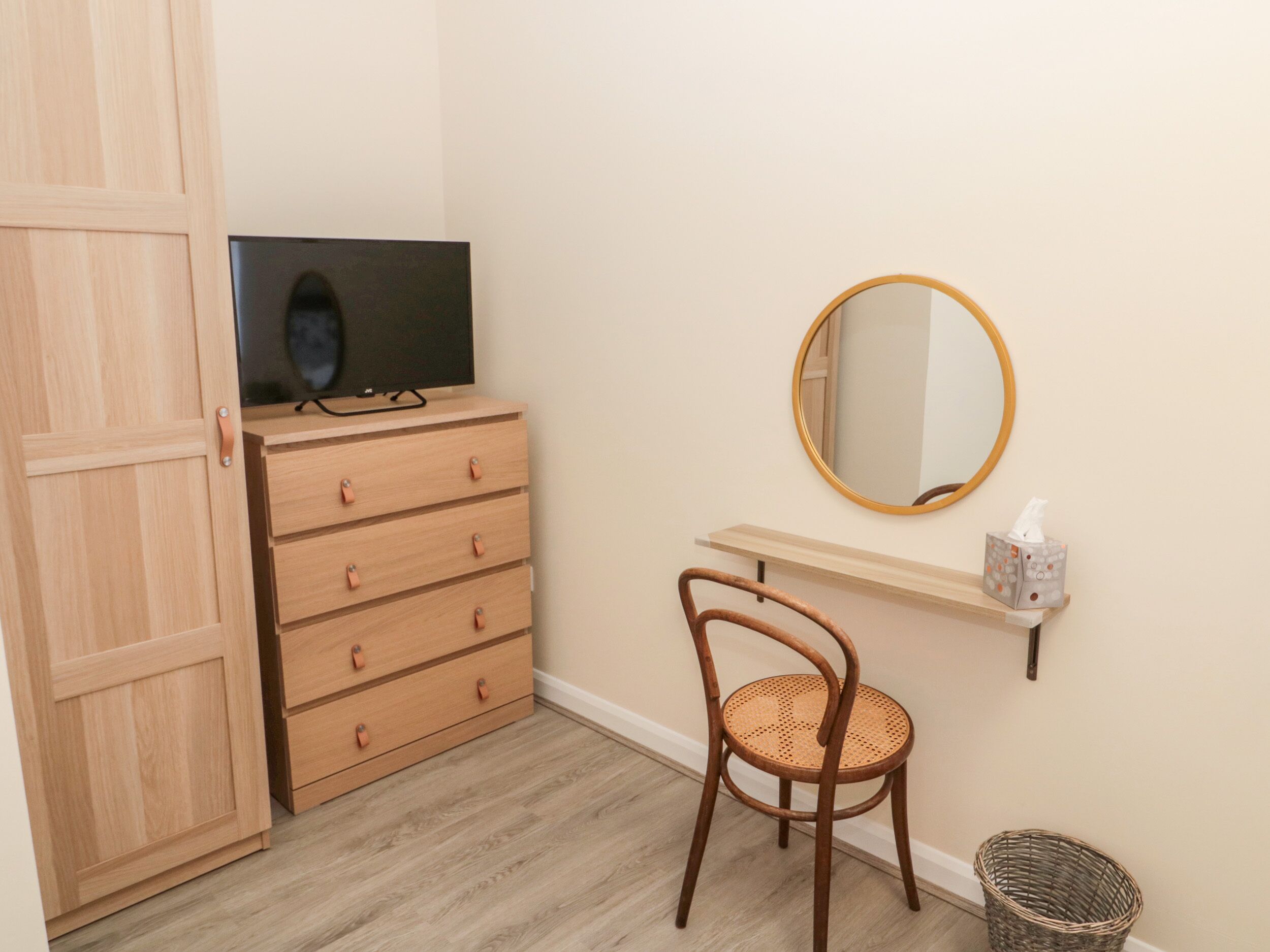 A room with a wooden wardrobe, a television on top, a chair, a mirror, and a small shelf with a tissue box.