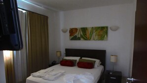 A hotel room with a bed, a nightstand, a lamp, a TV, and a window with curtains. - Lovely 2-bed Apartment in Santa Maria (Sal)