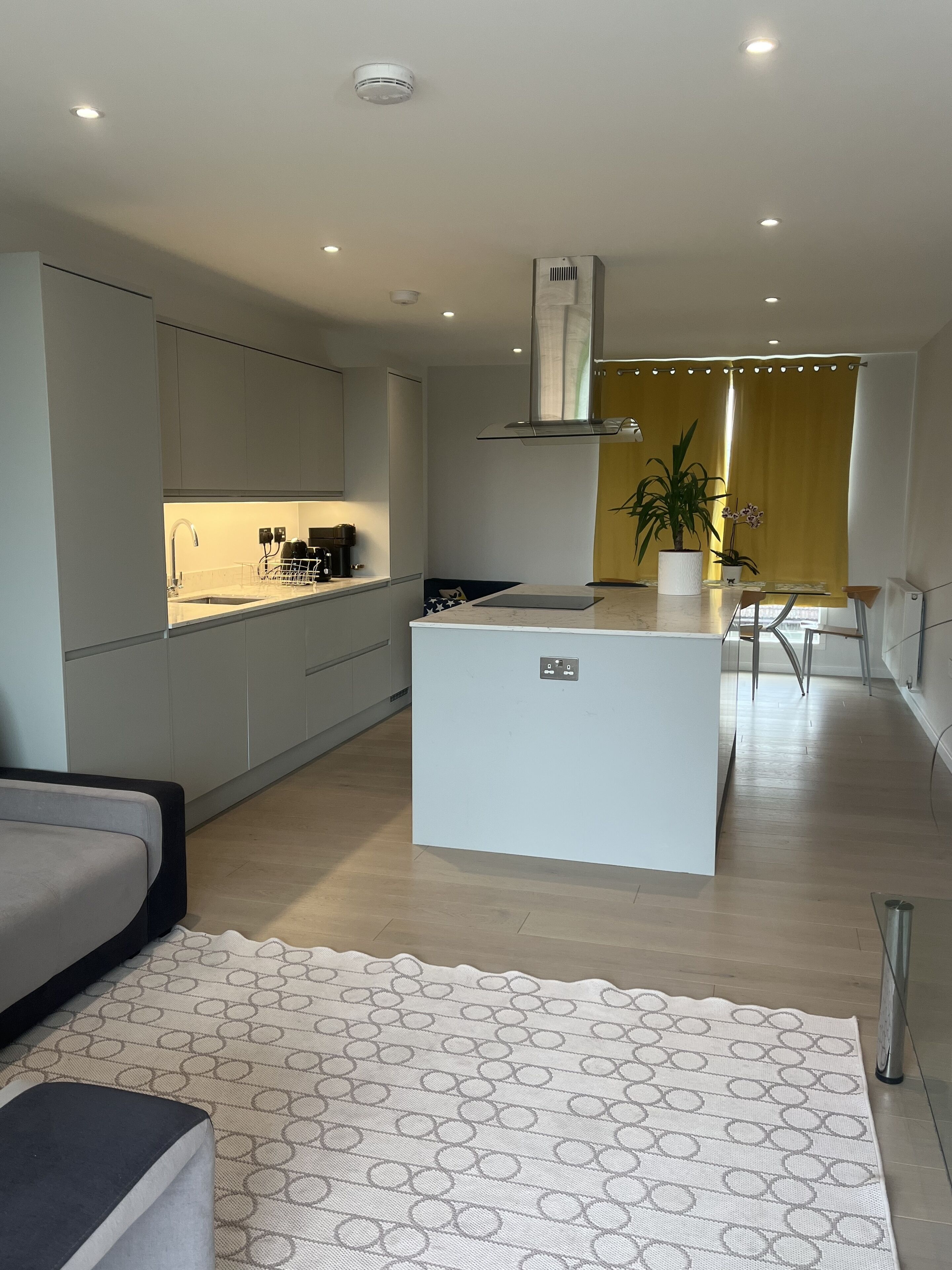 A modern kitchen with a central island, a dining area with yellow curtains, and a living space with a patterned rug.