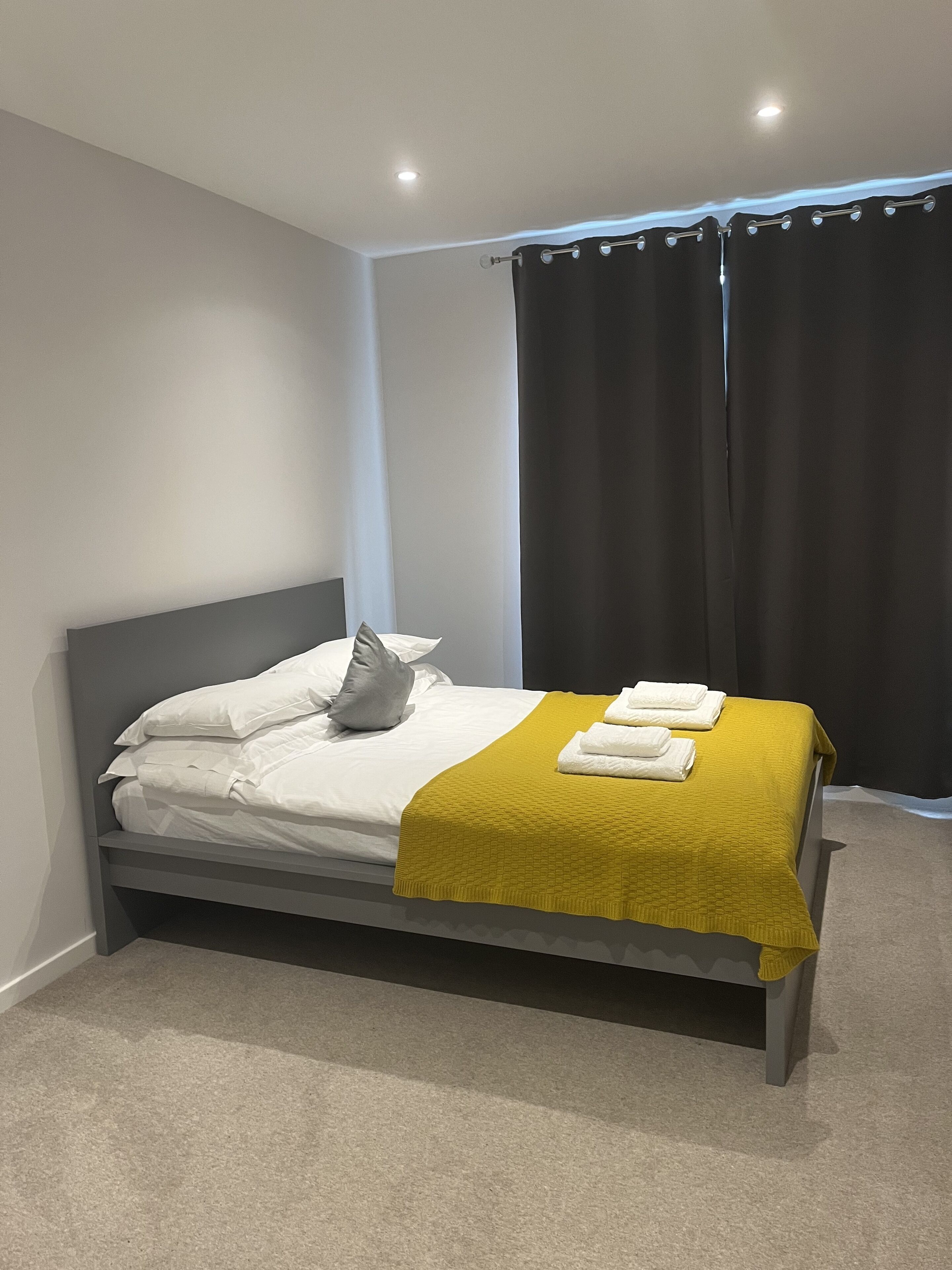 A neatly made bed with white linens and a mustard-colored blanket, a gray headboard, and a dark curtain to the right.