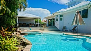 A swimming pool with a stone border, a covered patio with chairs, and a staircase leading to a covered area. - Mermaid's Crossing managed by Beach Retreats (Holmes Beach)