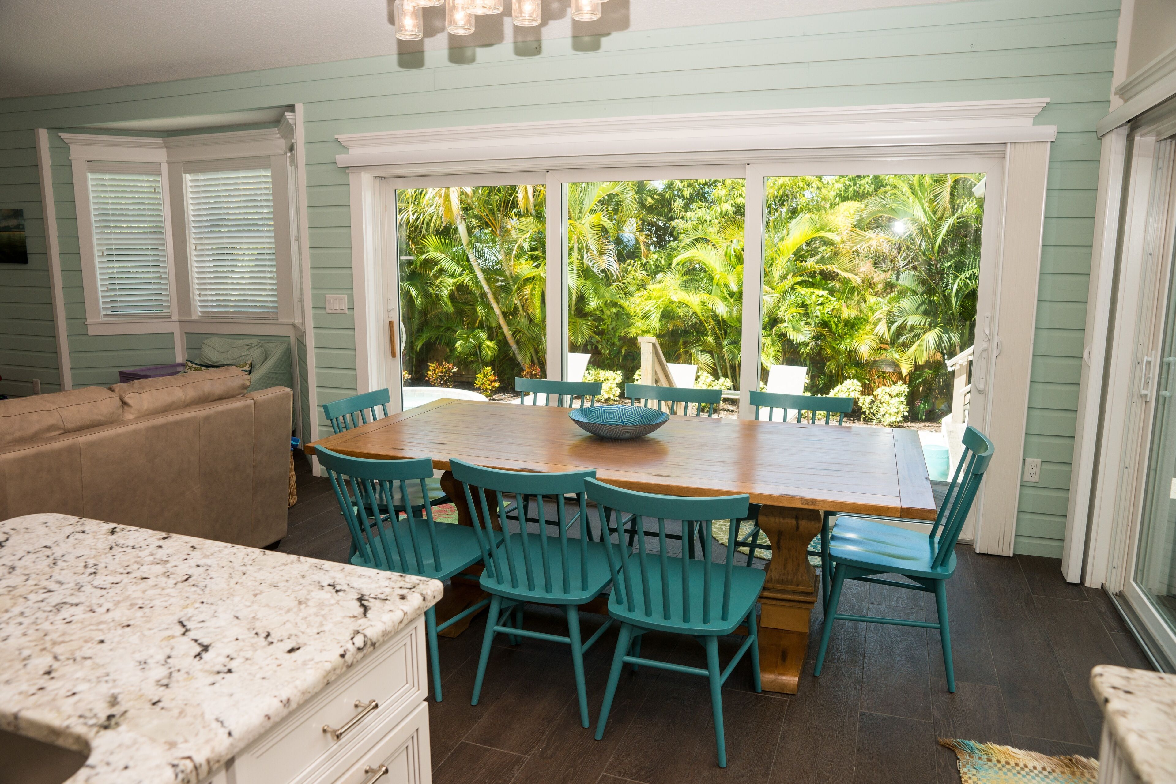 A dining area with a wooden table and teal chairs, a kitchen with a granite countertop, and a view of lush greenery through large windows.