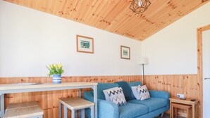Una habitación acogedora con un sofá azul, una mesa de comedor de madera y un taburete pequeño. Hay dos cuadros en la pared, una lámpara y una planta sobre la mesa.