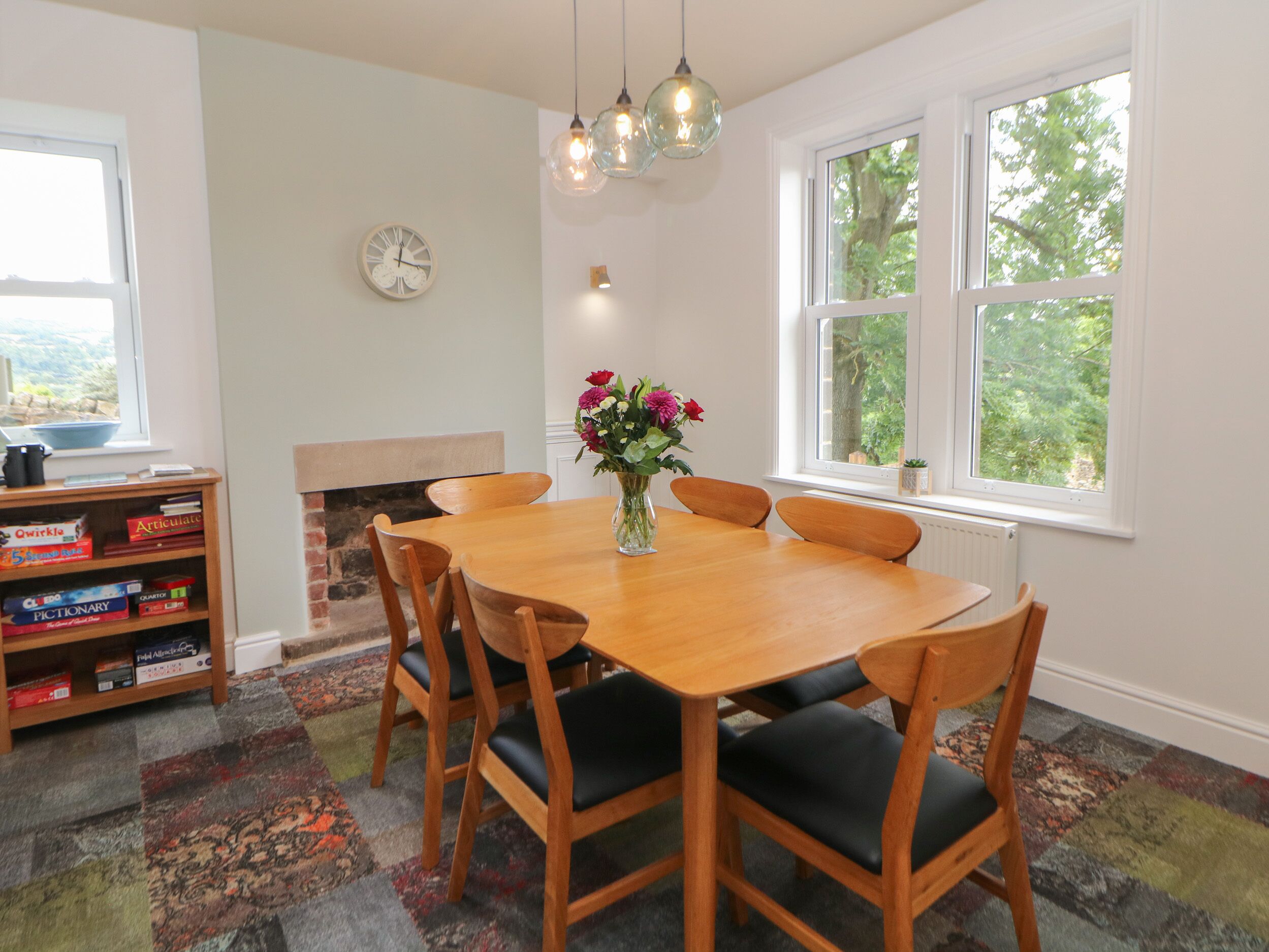 A dining room with a wooden table, six chairs, a vase of flowers, a bookshelf, a fireplace, and large windows with a view of trees.
