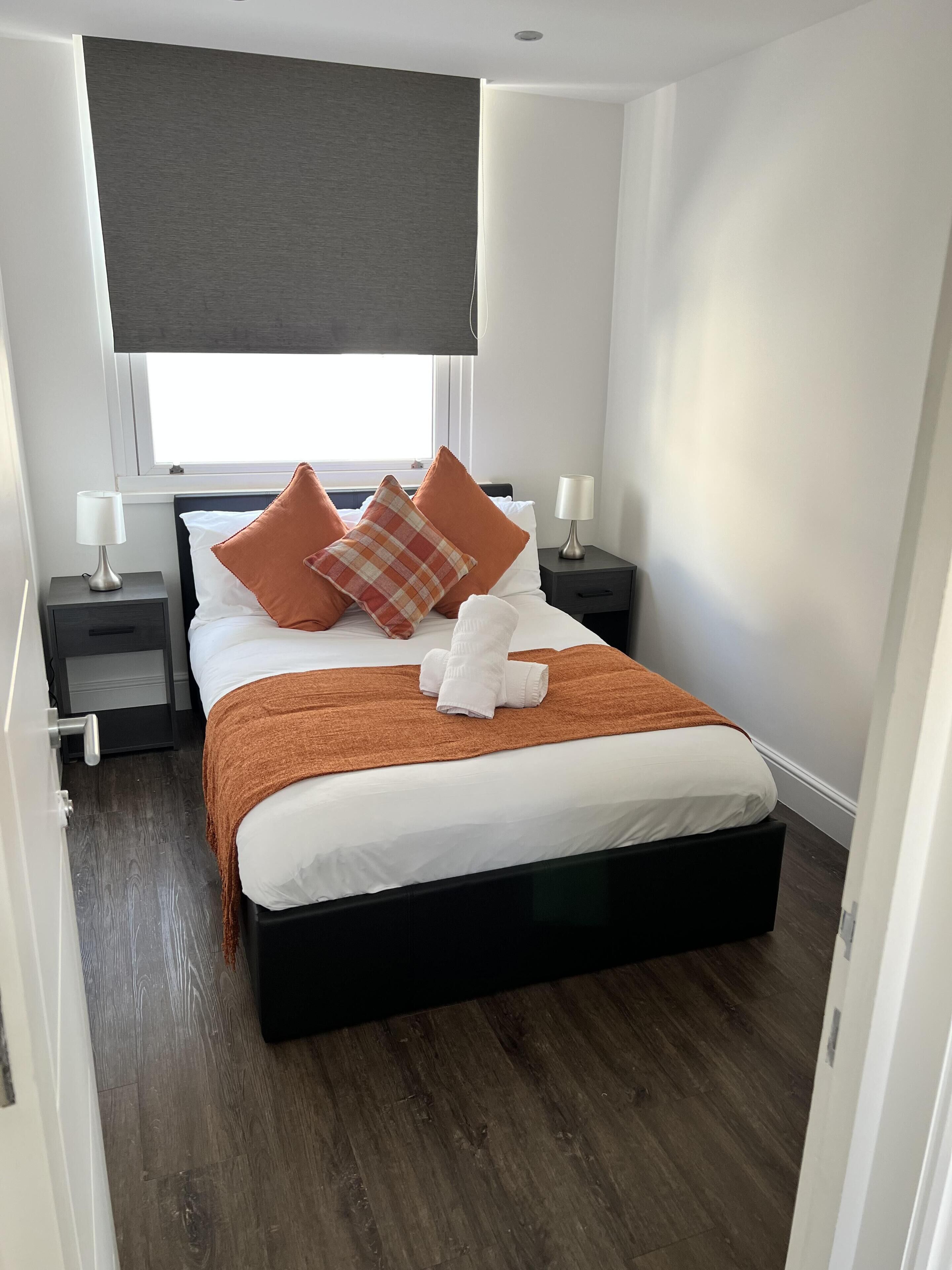 Une chambre à coucher bien rangée, avec un lit, deux tables de chevet, une fenêtre avec un store et un plancher en bois.