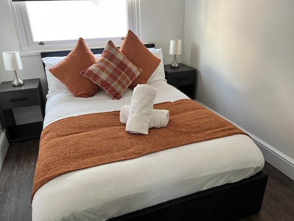Een net opgemaakt bed met witte lakens, een bruine plaid en een witte handdoek gevouwen op het bed. Er zijn twee nachtkastjes met lampen en een raam boven het bed.