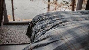 A bed with a plaid blanket, a window with a view of a lake and trees, and a wooden floor. - Luxurious Tree Top Cabin by the Lake (Iveland)