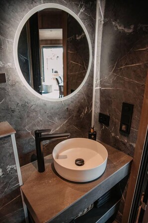 A modern bathroom with a round sink, a large mirror, and a marble wall. - Luxurious Tree Top Cabin by the Lake (Iveland)