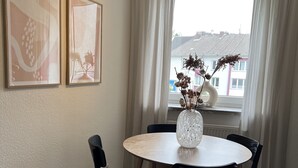 Un coin repas avec une table ronde, des chaises noires et un vase contenant des fleurs séchées. Deux tableaux encadrés ornent le mur.