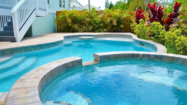 A swimming pool with a curved design, surrounded by a stone deck and lush greenery. - Turtle's Bay managed by Beach Retreats (Holmes Beach)