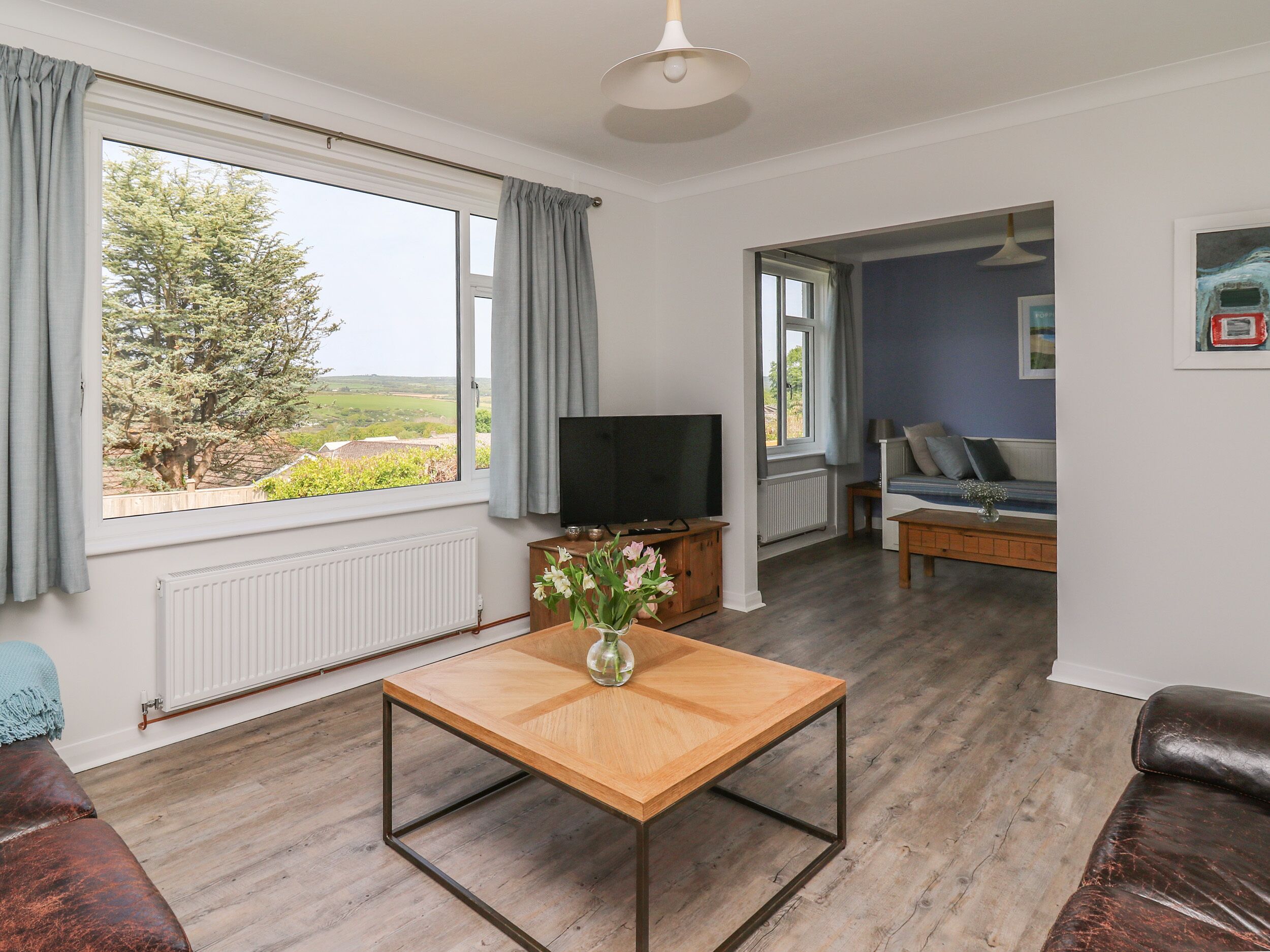Una sala de estar con una mesa de centro de madera, un televisor y un sofá. Hay una ventana con cortinas y vistas a árboles.