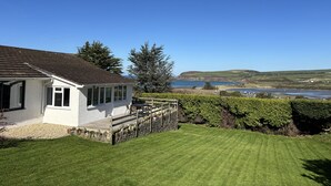 Une maison blanche avec des fondations en pierre, une terrasse en bois et une vue sur la mer et les collines.
