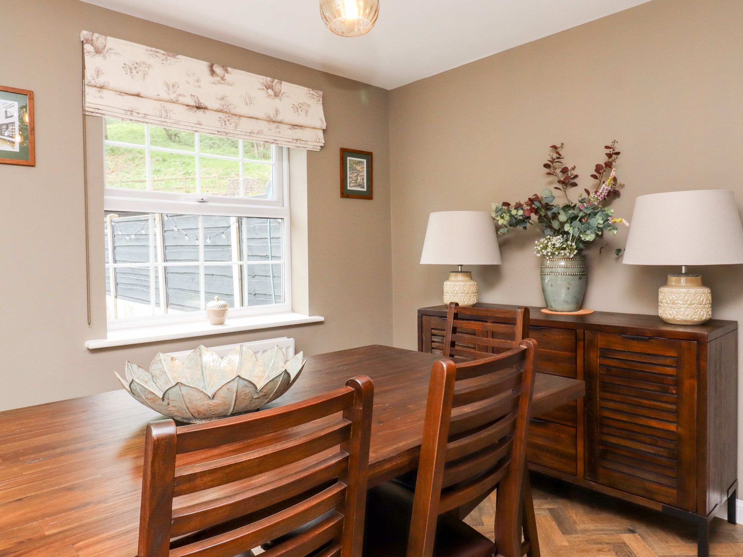 A dining room with a wooden table, chairs, and a window with a view of the outside. There are two lamps on the table and a vase with flowers on the sideboard.