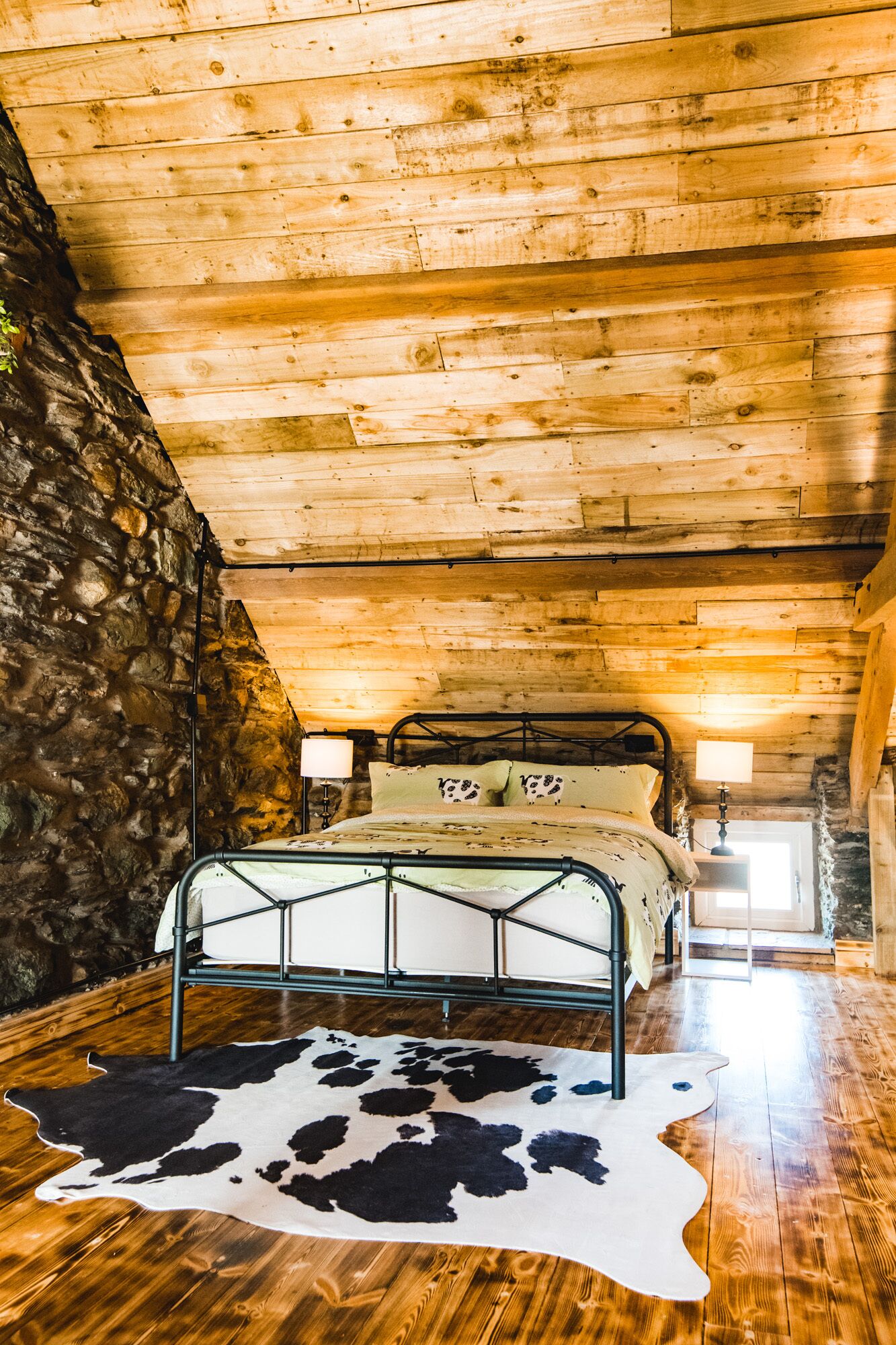 A rustic bedroom with a stone wall, a metal bed frame, and a cowhide rug.