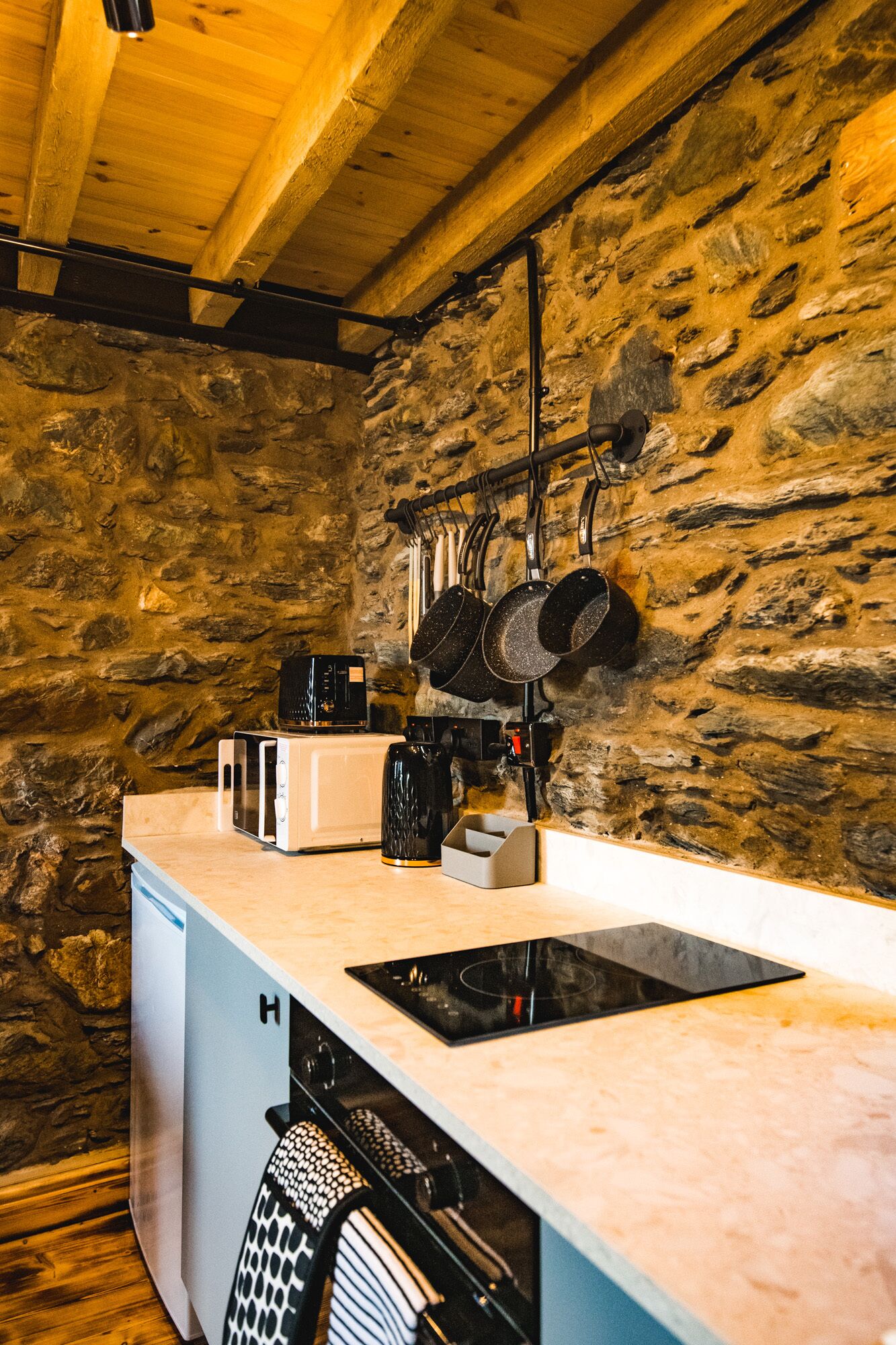 A compact kitchen with a stone wall, a built-in microwave, a toaster, a kettle, and a hanging pot rack.