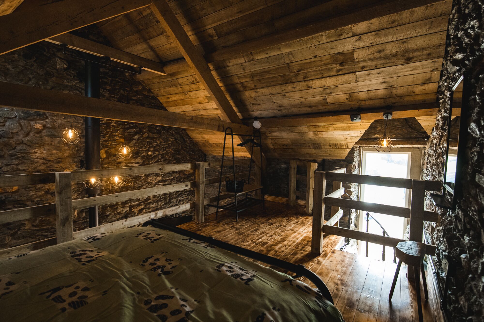 Ein rustikales Schlafzimmer im Dachgeschoss mit schrägen Wänden, freiliegenden Holzbalken, einer Steinwand und einem Bett mit gemustertem Überwurf.