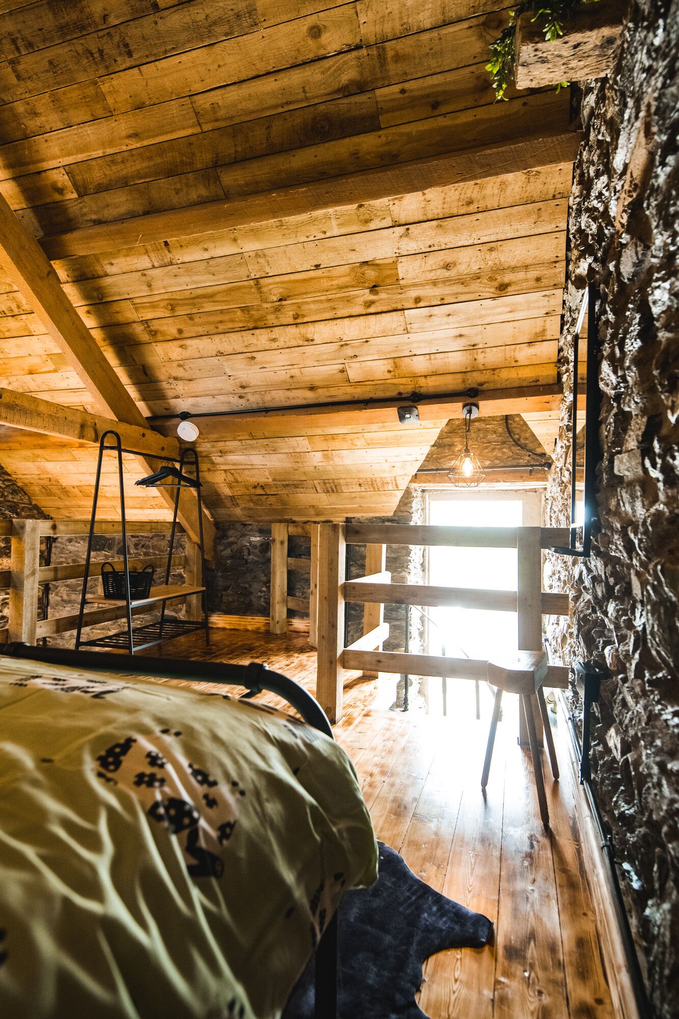 A rustic attic bedroom with exposed wooden beams, a stone wall, and a wooden ladder.