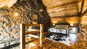 Ein rustikales Schlafzimmer mit Steinwand, Holzbalkendecke und einem Bett mit Kuhfellteppich.