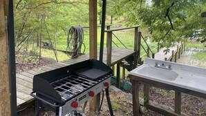 A covered outdoor area with a grill and sink, surrounded by wooden structures and greenery. - Lake Wateree cottage (Ridgeway)