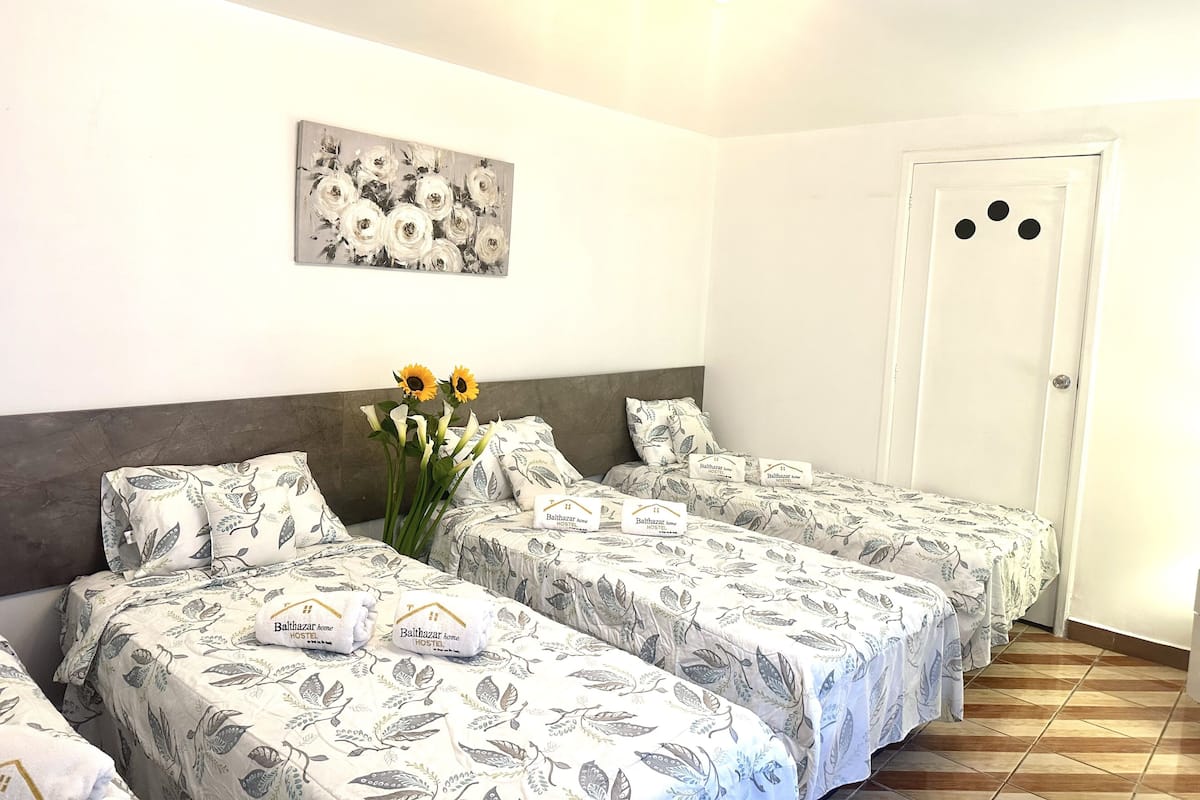 A hotel room with three beds, each with a floral-patterned bedspread, a painting of flowers on the wall, and a vase with sunflowers on the nightstand.