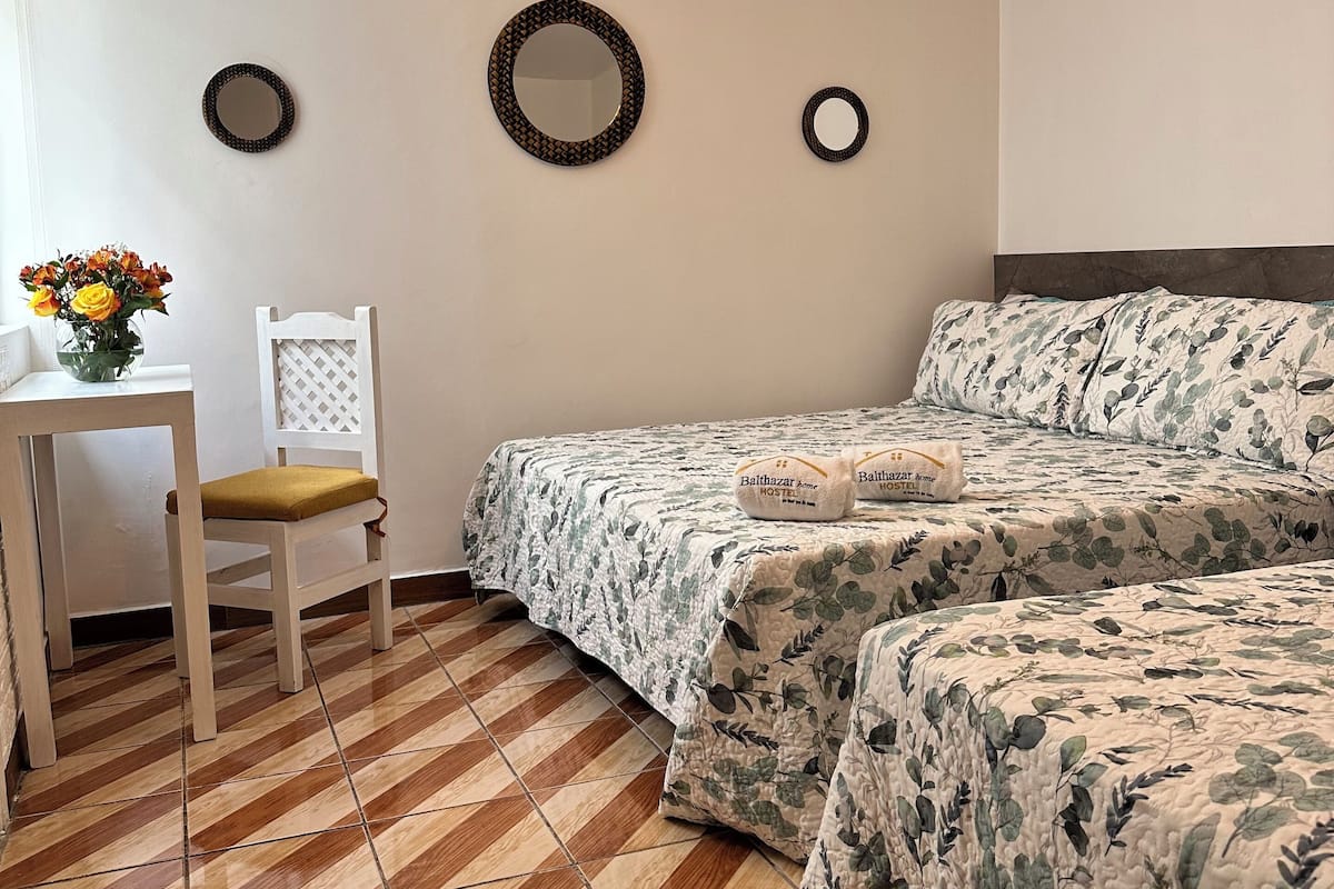A room with a bed, a chair, a small table with a vase of flowers, and two wall-mounted mirrors.