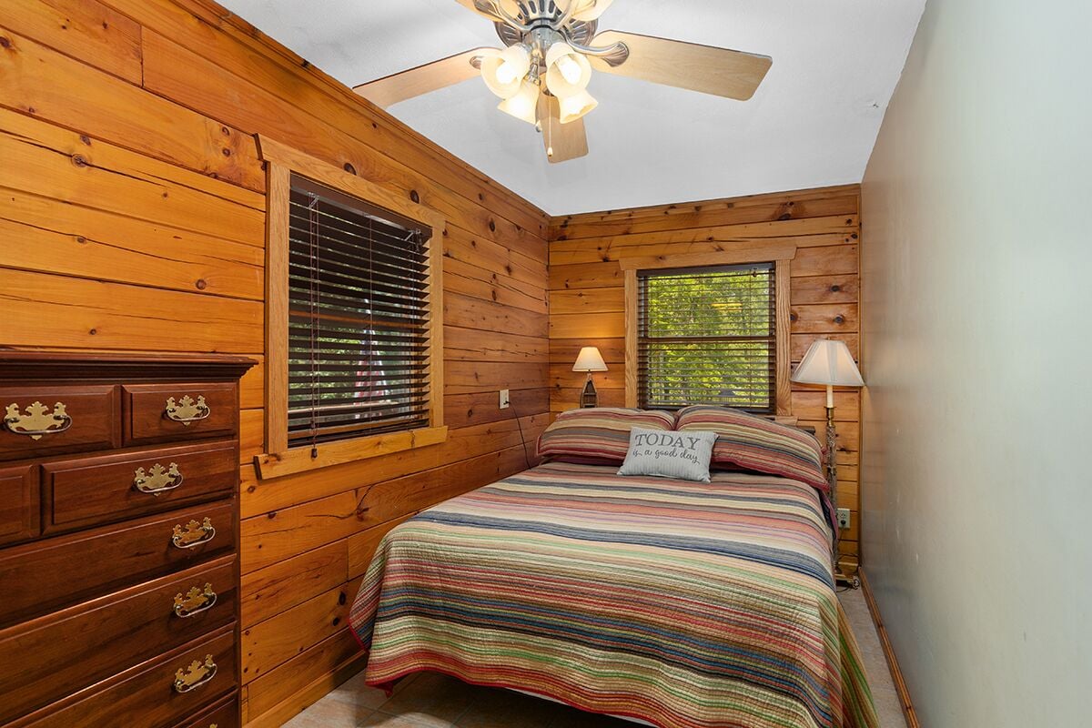 Un dormitorio de cabaña de madera con cama, cómoda, dos ventanas, ventilador de techo y lámparas.